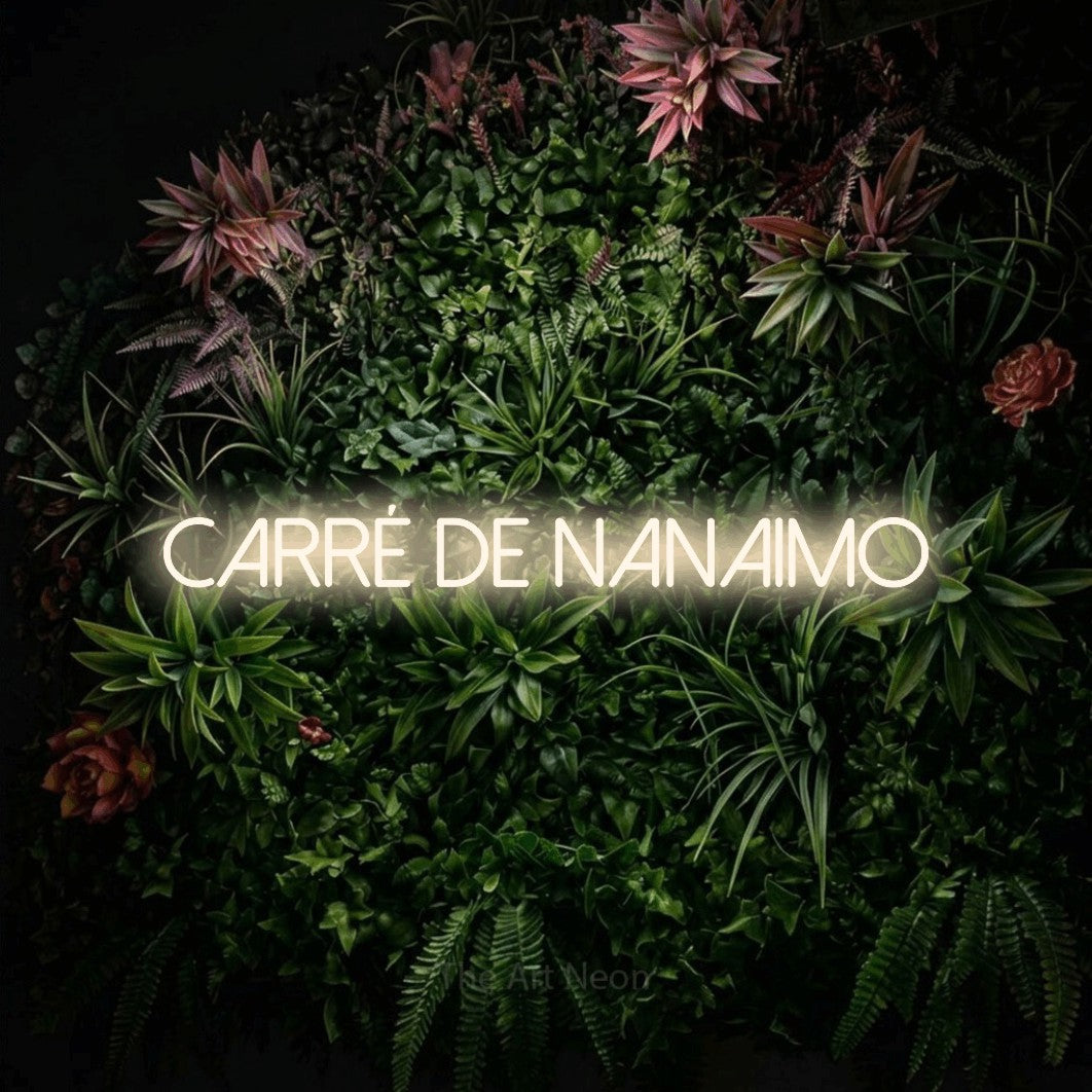 Décoration murale en néon LED « Carré de Nanaimo » en blanc chaud, en lettres fines sans empattement, installée sur un mur végétal fleuri, pour un décor canadien.