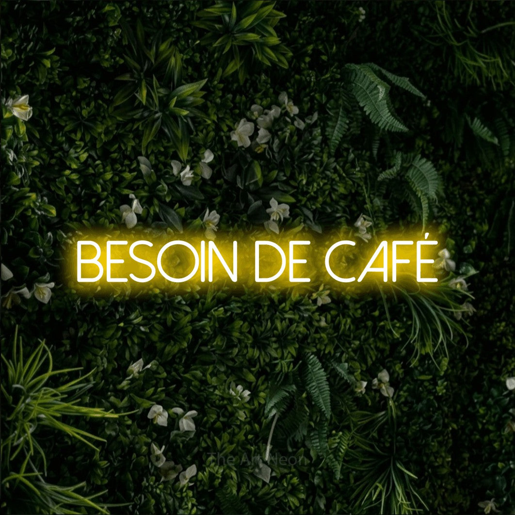 “Besoin de café” LED neon sign in yellow, with thin sans-serif lettering, shown against a dense green plant wall.