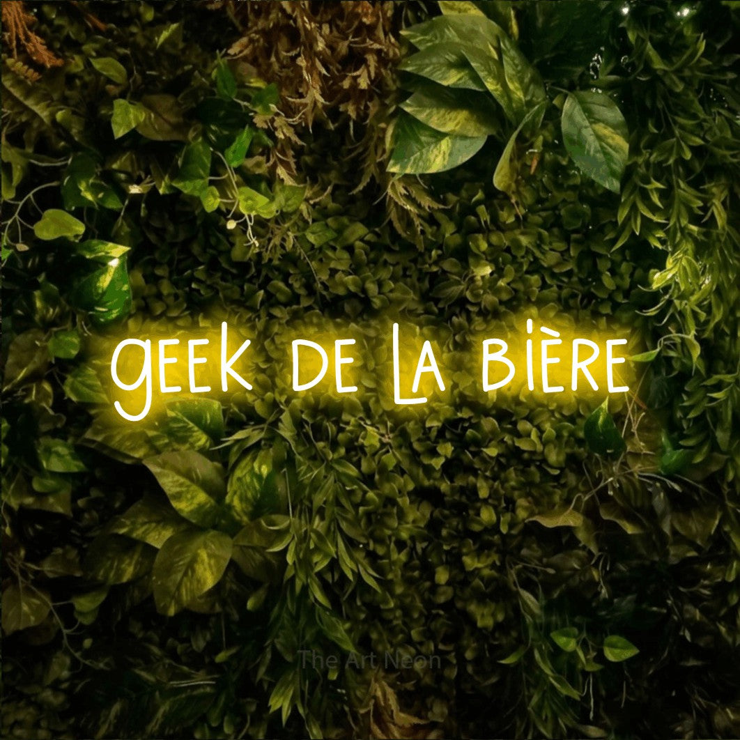 “Geek de la bière” LED neon sign in yellow, with narrow hand-drawn lettering, shown against bright green and yellow foliage.