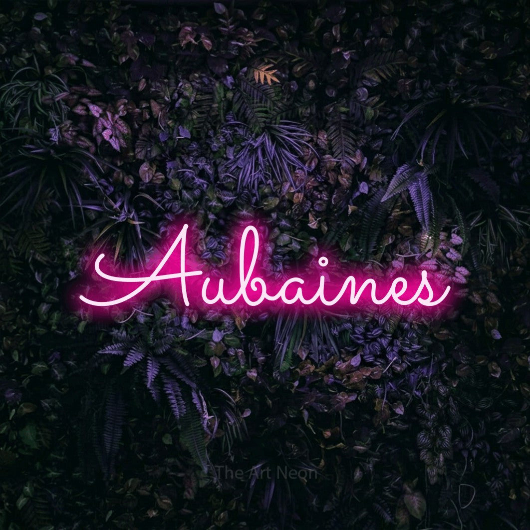 “Aubaines” LED neon sign in hot pink, with looped cursive lettering, shown against a dark wall with purple flowers.