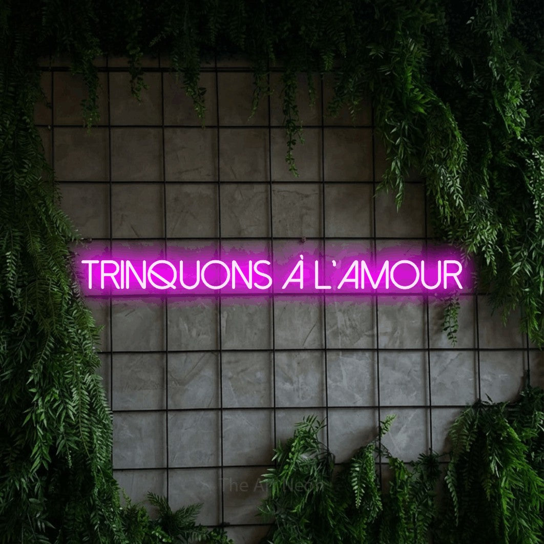 “Trinquons à l'amour” LED neon sign in pink, with thin sans-serif lettering, shown against an ivy-covered concrete grid.