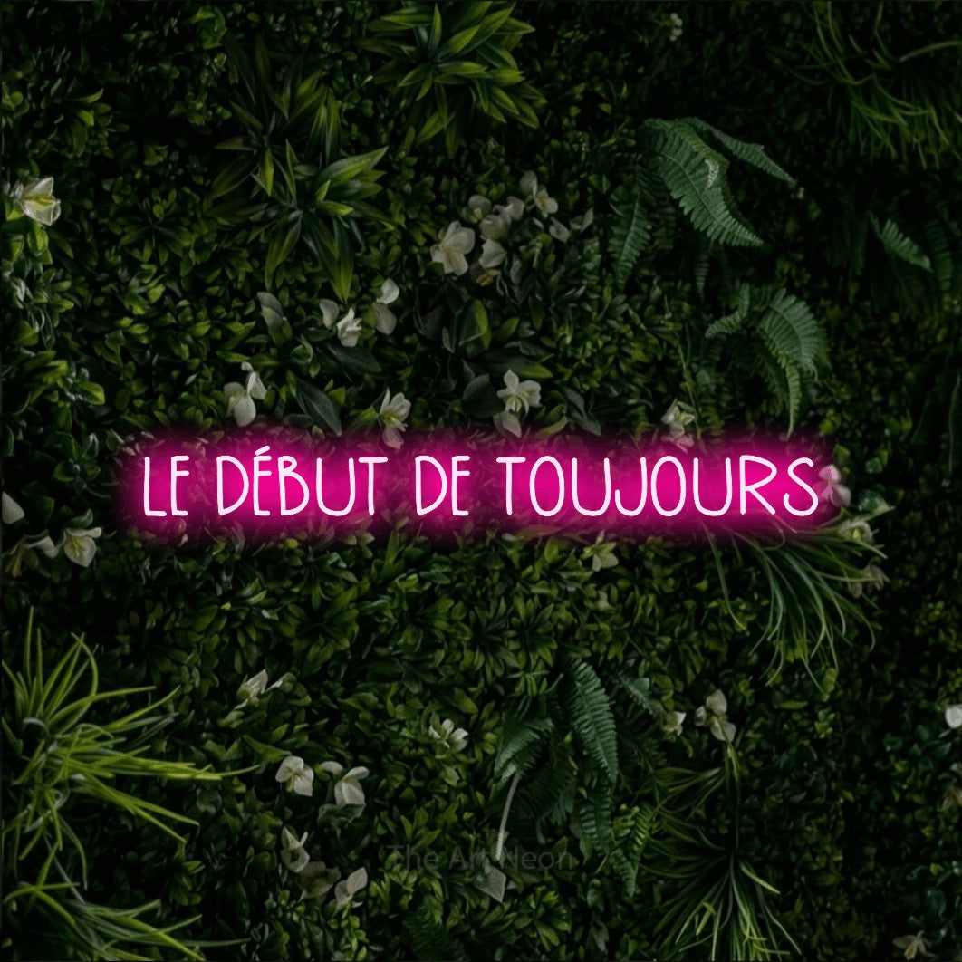 “Le début de toujours” LED neon sign in hot pink, with narrow hand-drawn lettering, shown against a dense green plant wall.