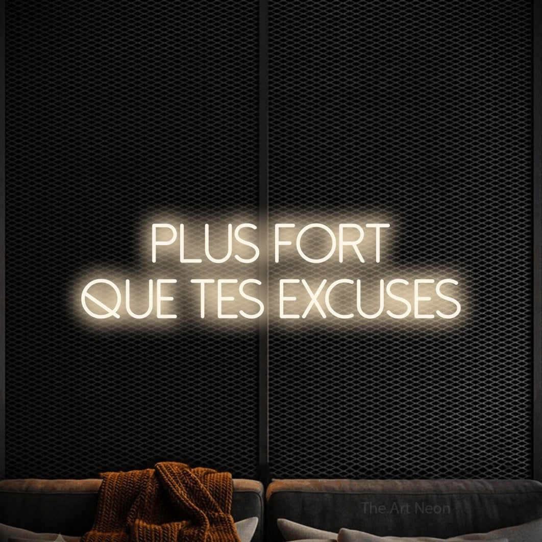 Néon LED « Plus fort que tes excuses » sur deux lignes en blanc chaud, en lettres fines sans empattement, mis en scène dans un coin salon moderne aux tons noirs, pour un espace d’entraînement.