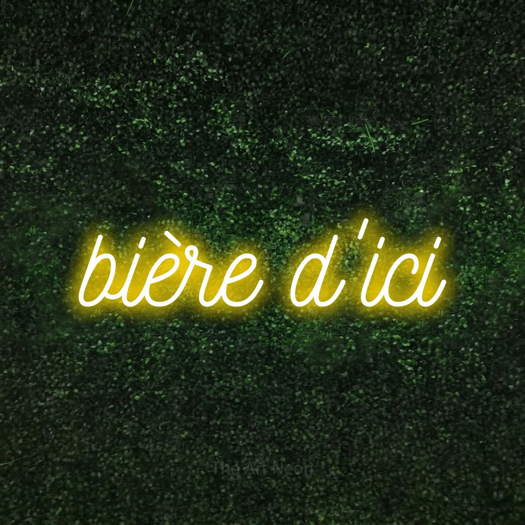 “Bière d'ici” LED neon sign in yellow, with ornate cursive lettering, shown against a textured green hedge.