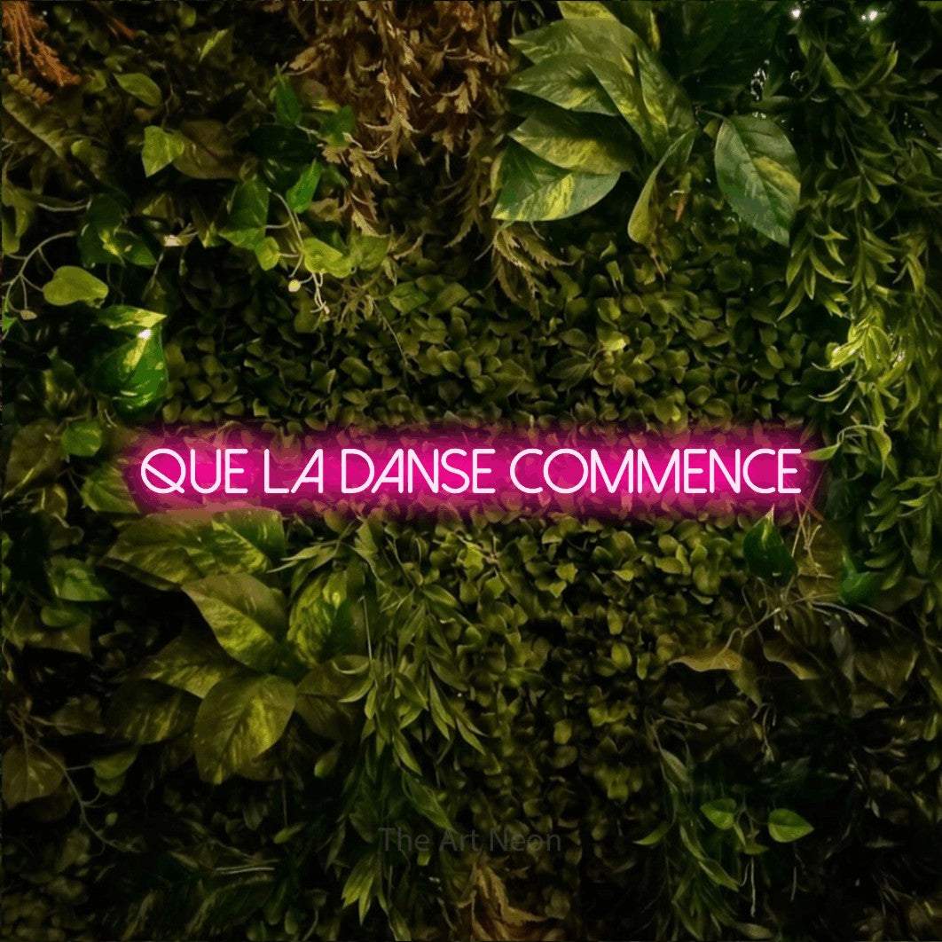 “Que la danse commence” LED neon sign in hot pink, with thin sans-serif lettering, shown against bright green and yellow foliage.