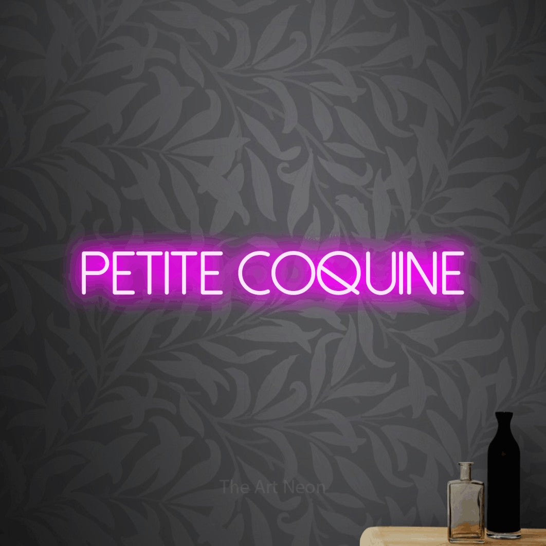 Aperçu d’une enseigne néon LED « Petite coquine » en rose, en lettres fines sans empattement, photographiée sur un papier peint botanique gris, pour une chambre d’enfant.