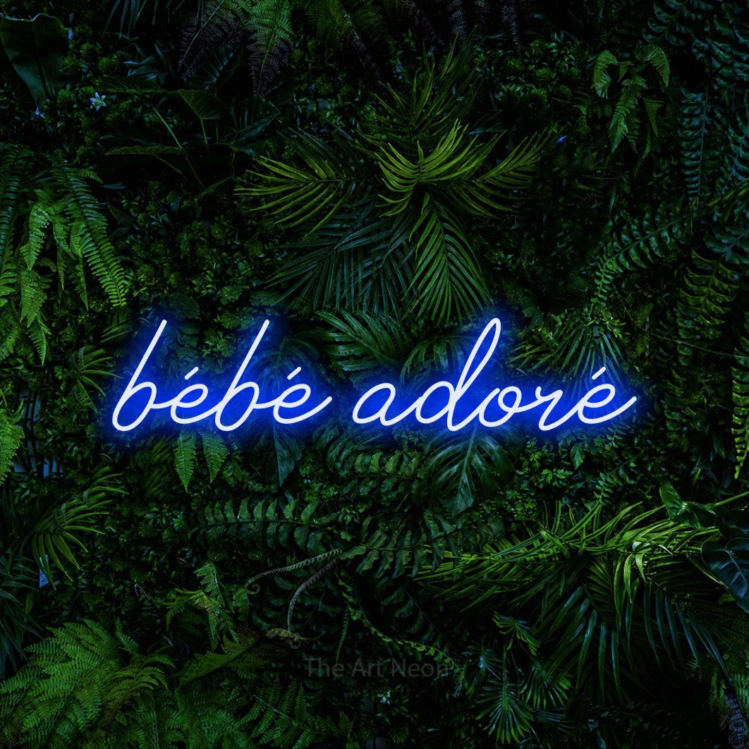 “Bébé adoré” LED neon sign in blue, with cursive lettering, shown against dense tropical foliage.